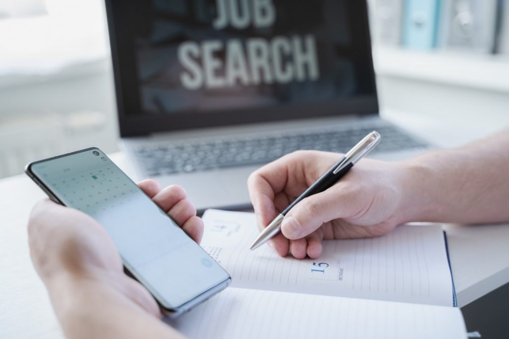 job seeker plotting schedule and planning for his job search to land a job on his notebook while using his phone to check calendar and laptop to look for job search opportunities